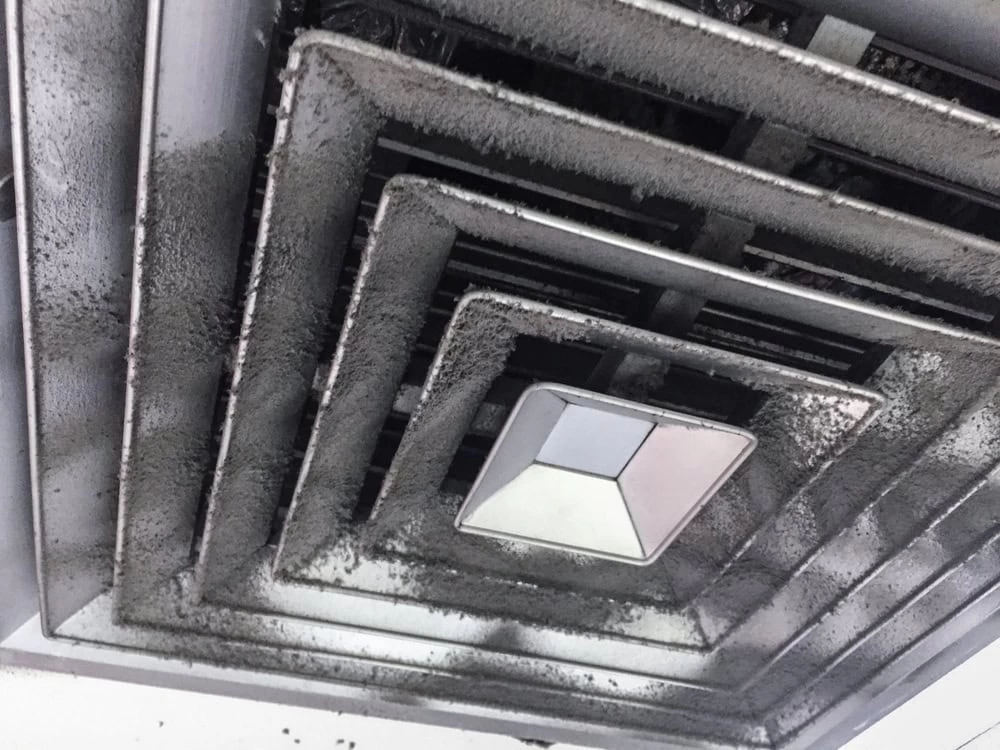 A close-up of a dirty, dust-covered air vent on a ceiling, with layers of grime on the vent slats and a light fixture in the center&mdash;highlighting the need for professional air duct services New Jersey.