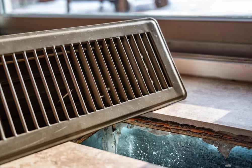 A close-up of a vent cover removed from a floor vent, revealing ductwork below with sunlight streaming in—a scene often encountered during air duct services New Jersey.