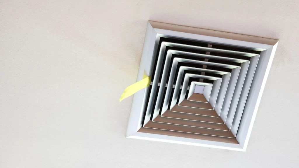 A yellow sticky note is caught on a square ceiling air vent with angled slats, mounted on a white ceiling&mdash;reminding you it may be time to book air duct services New Jersey for cleaner indoor air.