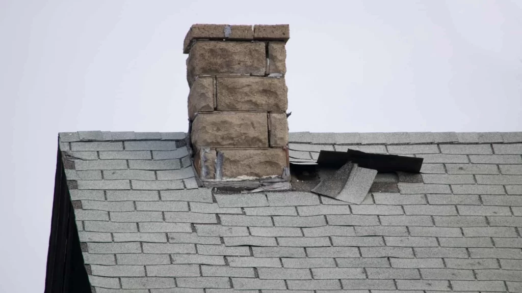 A brick chimney on a gray shingled roof with visible damage; several shingles are lifted and displaced near the chimney, exposing the underlying roofing materials&mdash;issues that may also affect air duct services New Jersey homeowners rely on.