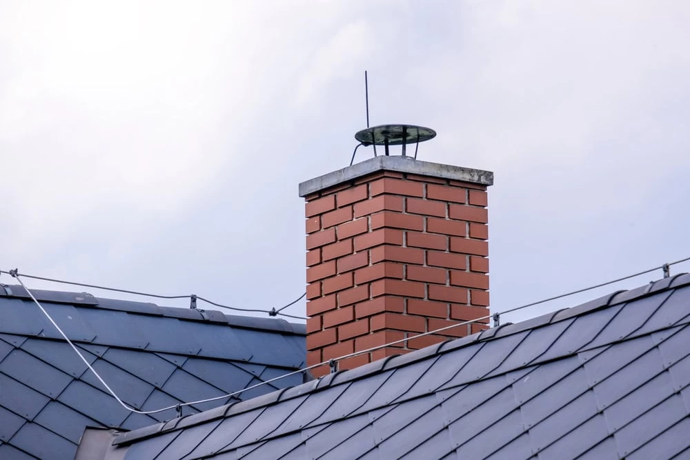 A brick chimney with a metal cap rises above a gray metal roof against a cloudy sky. Thin wires are attached along the roof&rsquo;s edge, possibly for lightning protection&mdash;a common sight during air duct services New Jersey homeowners schedule.