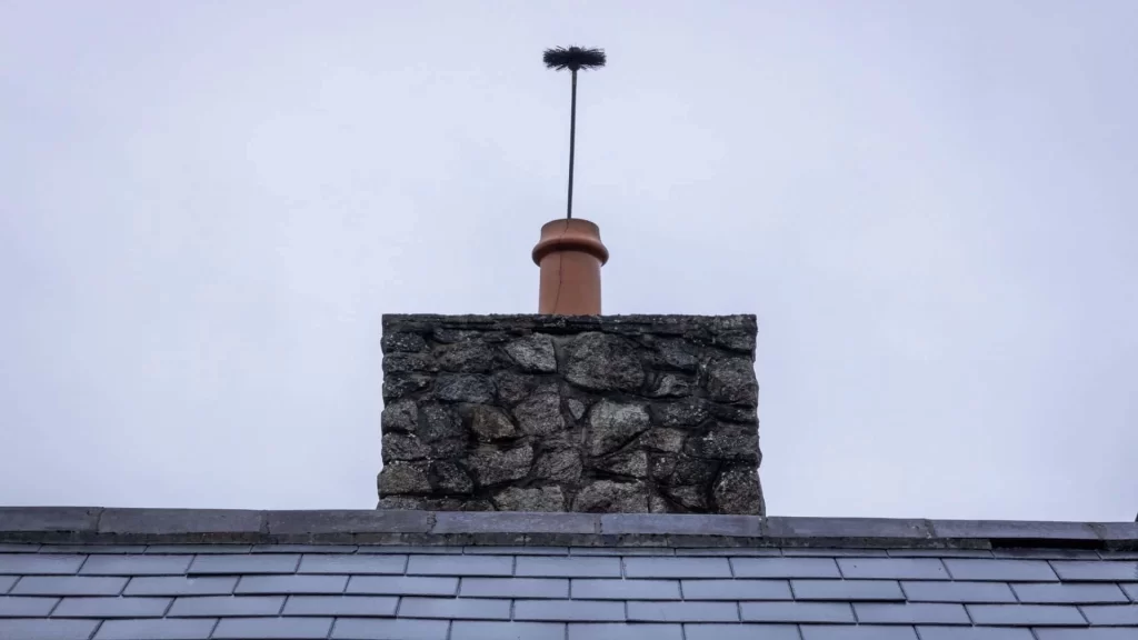 A stone chimney with a terracotta pipe and chimney brush atop, set against a cloudy, gray sky, viewed from behind a slate-tiled roof&mdash;a scene evoking the need for expert dryer vent cleaning New Jersey homeowners trust.