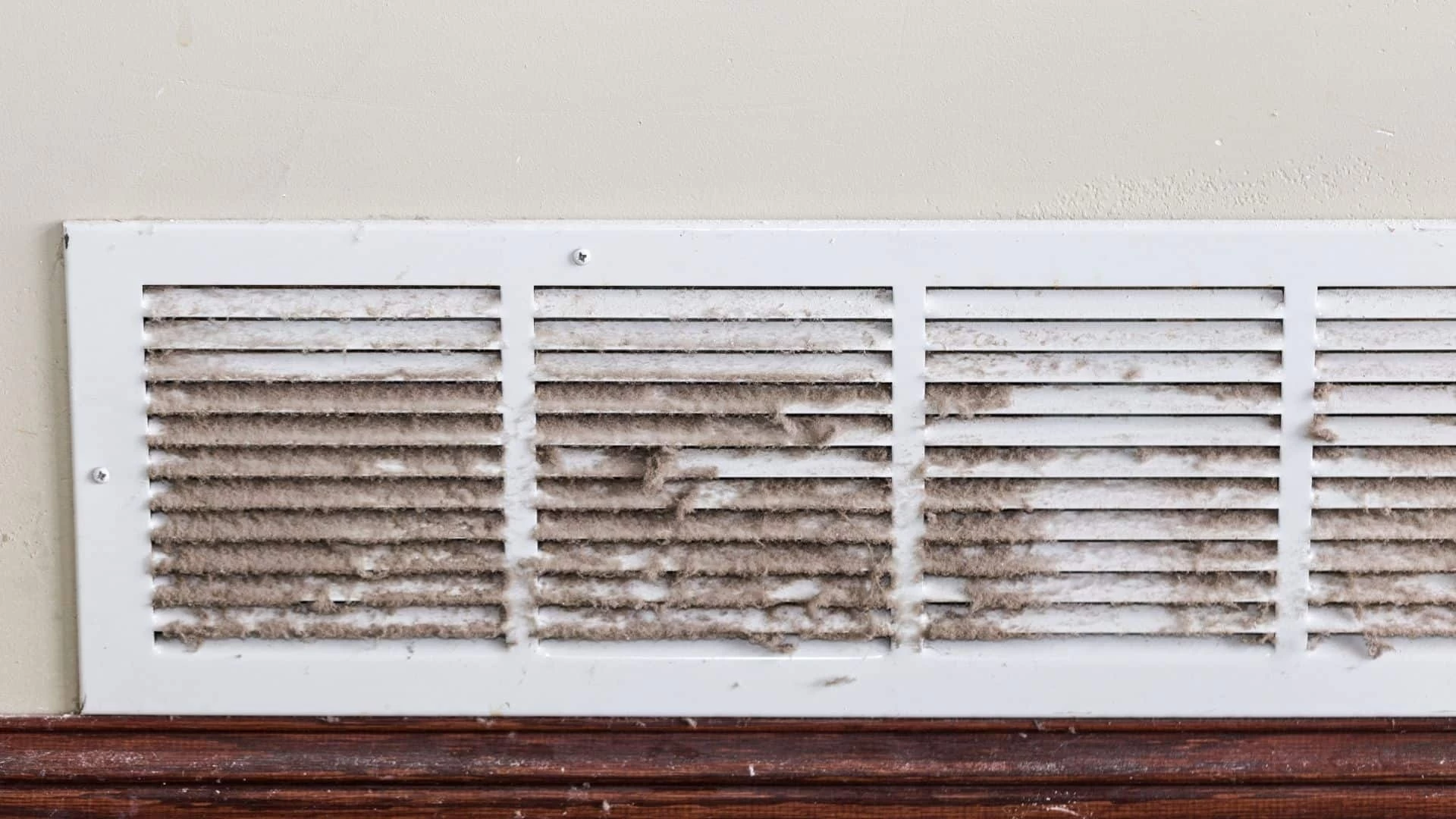 A white air vent covered in thick dust and dirt is mounted on a beige wall above a wooden baseboard, highlighting the need for professional air duct services New Jersey.