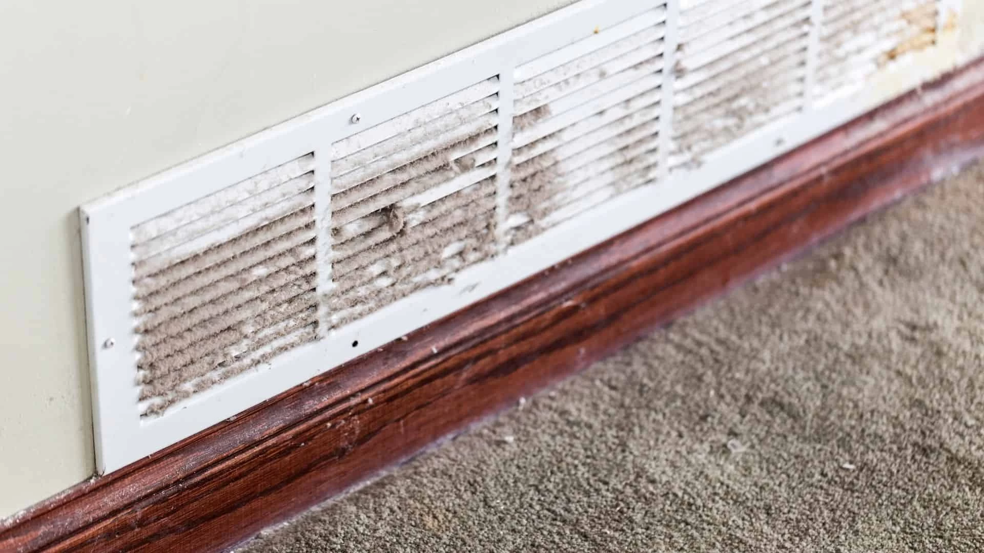 A white wall vent covered in dust and dirt is mounted above a brown wooden baseboard and gray carpeted floor, highlighting the need for professional air duct cleaning services New Jersey.