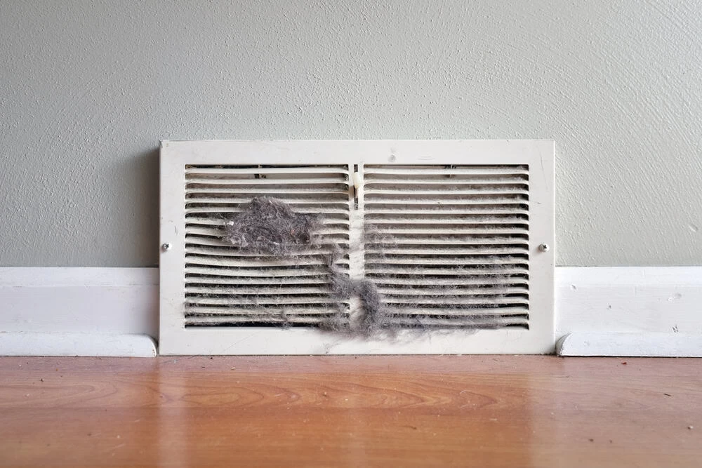 A white floor vent cover against a gray wall and baseboard shows dust and lint buildup on the slats above a wooden floor&mdash;a clear sign it's time to consider air duct services New Jersey for a cleaner, healthier home.