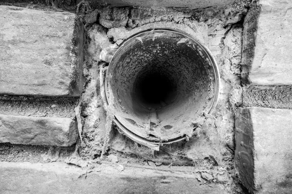 A close-up, black-and-white photo of a round, empty pipe embedded in a rough brick wall, surrounded by cobwebs and dust&mdash;an evocative reminder of the importance of dryer vent cleaning New Jersey.