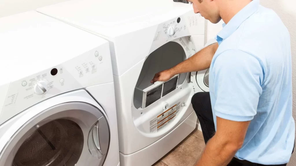 A man in a light blue shirt kneels in front of a white clothes dryer, removing the lint trap for cleaning. Just like dryer vent cleaning New Jersey recommends, he keeps his laundry area&mdash;complete with a matching washer&mdash;safe and efficient.