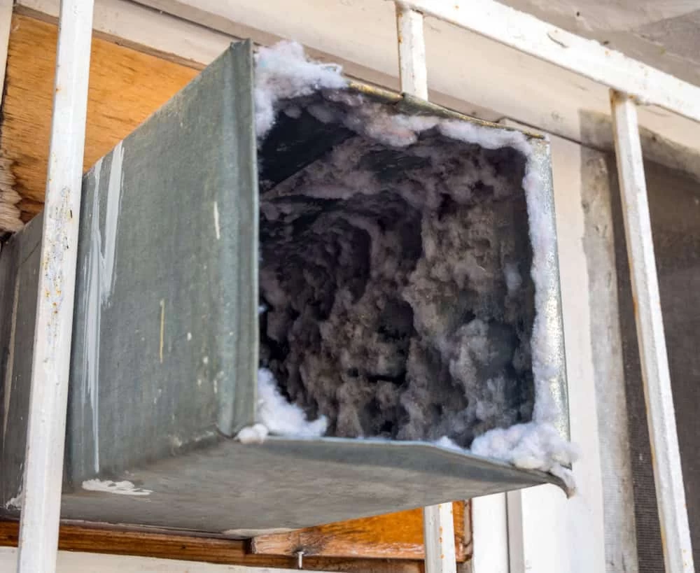 A close-up of a metal air duct vent with a large buildup of dust and lint inside, indicating it needs dryer vent cleaning New Jersey. The vent is attached to an exterior wall with white bars.