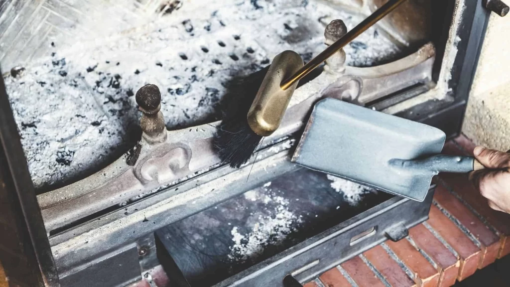 A person cleaning out ashes from a fireplace using a metal dustpan and brush, with gray ash and soot visible inside the fireplace&mdash;reminding homeowners of the importance of regular dryer vent cleaning New Jersey for a safer, cleaner home.