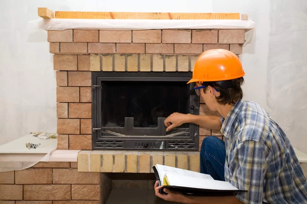 A person wearing an orange hard hat and plaid shirt inspects a brick fireplace, pointing at the glass door while holding an open notebook&mdash;demonstrating the attention to detail found in top air duct services New Jersey relies on.