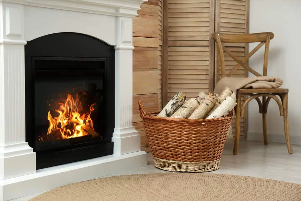 A cozy indoor scene with a lit fireplace, a wicker basket of firewood, a wooden chair draped with a folded blanket, and a round woven rug&mdash;perfect for relaxing after expert dryer vent cleaning New Jersey has to offer.