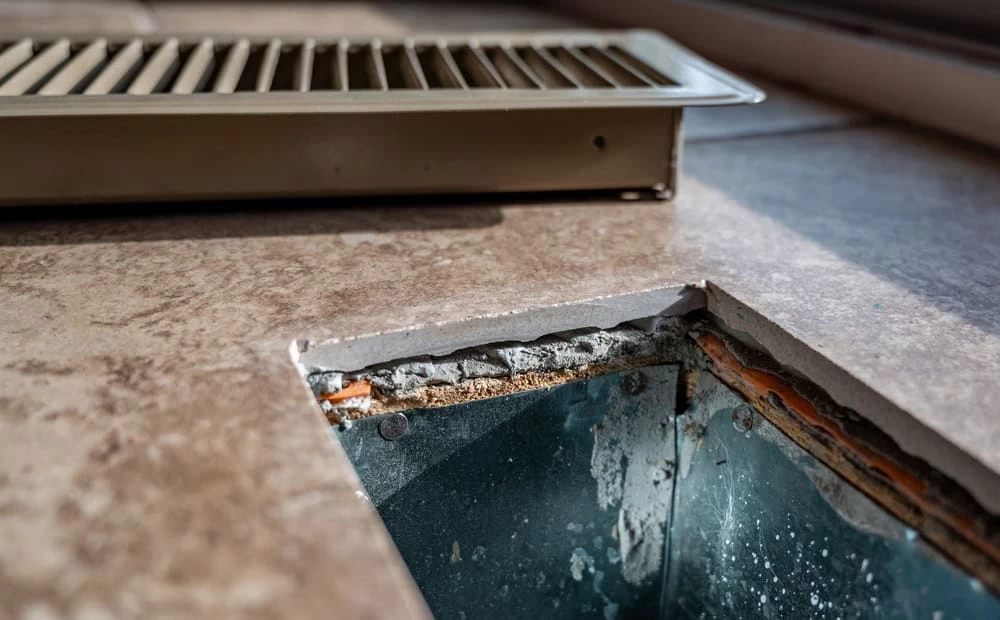 A close-up of a floor vent opening with the rectangular metal vent cover removed, exposing the ductwork and rough edges of the surrounding floor&mdash;ideal for highlighting air duct services New Jersey. The vent cover rests nearby on the tiled floor.