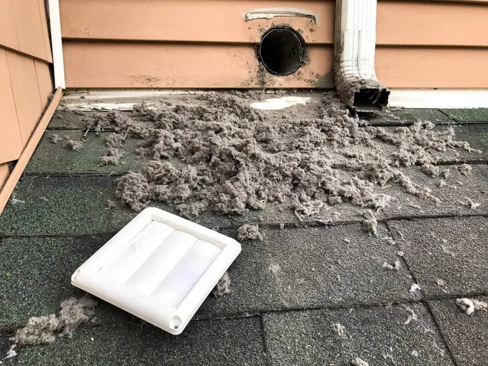 A detached dryer vent cover lies on a shingled roof, surrounded by gray lint from an exposed vent opening&mdash;highlighting the need for professional dryer vent cleaning New Jersey homeowners can trust.