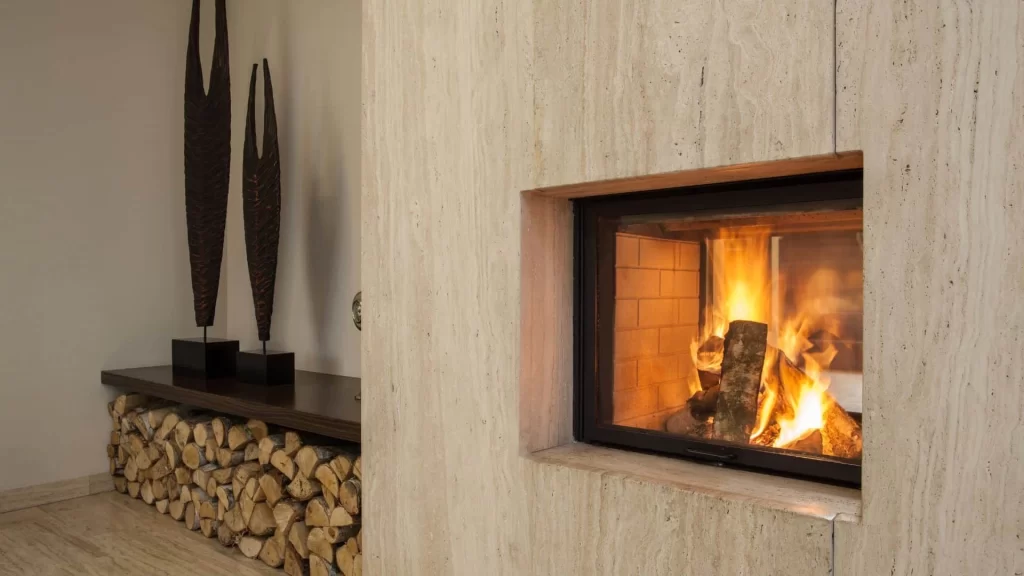 A modern fireplace with a glass front and burning logs, set in a light stone wall. Stacked firewood sits beneath a dark wooden shelf with two tall sculptures&mdash;perfect comfort after professional dryer vent cleaning New Jersey.