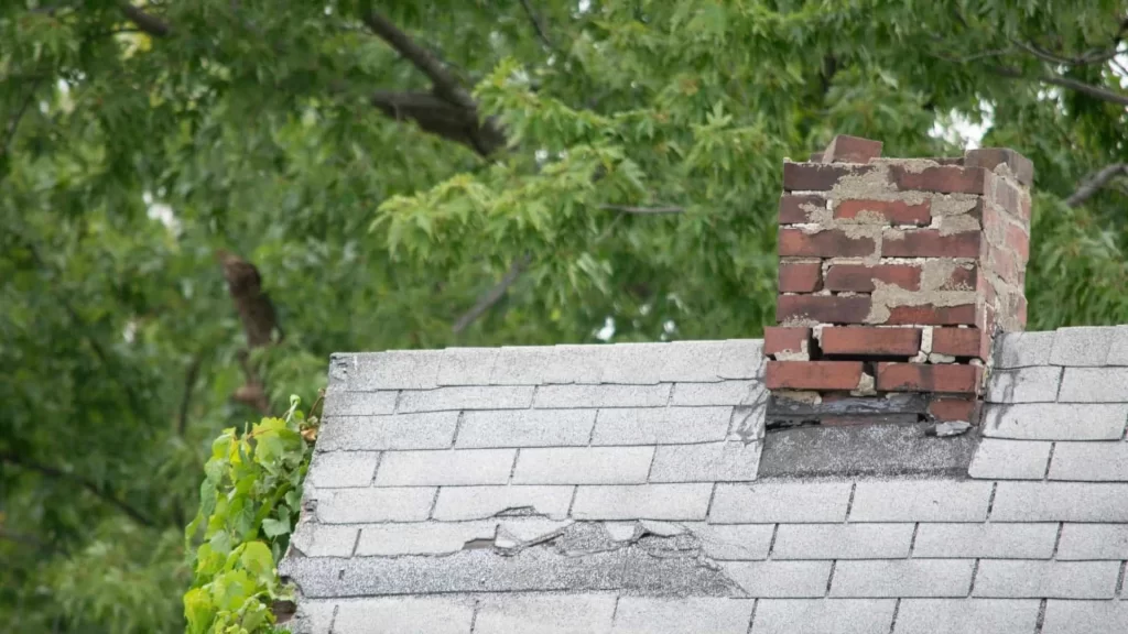 A weathered brick chimney rises above a shingle-covered roof with missing and cracked tiles, while green leafy trees frame the scene&mdash;an ideal setting to consider air duct services New Jersey for improved home safety.