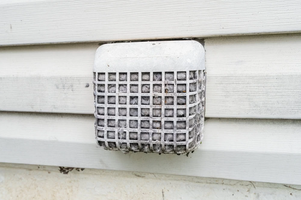 A white vent cover on an exterior wall is clogged with gray lint and debris, showing it needs cleaning&mdash;a clear reminder of the importance of regular dryer vent cleaning New Jersey homeowners should consider.