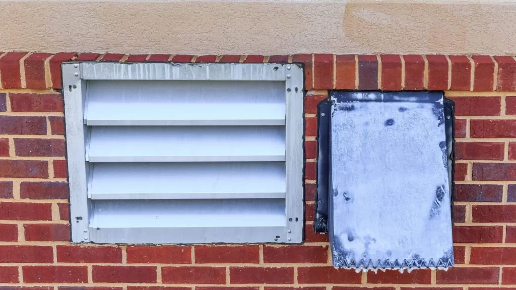 A red brick wall with a beige trim features a silver metal vent&mdash;ideal for dryer vent cleaning New Jersey experts&mdash;on the left and a frosted or weathered rectangular panel on the right.