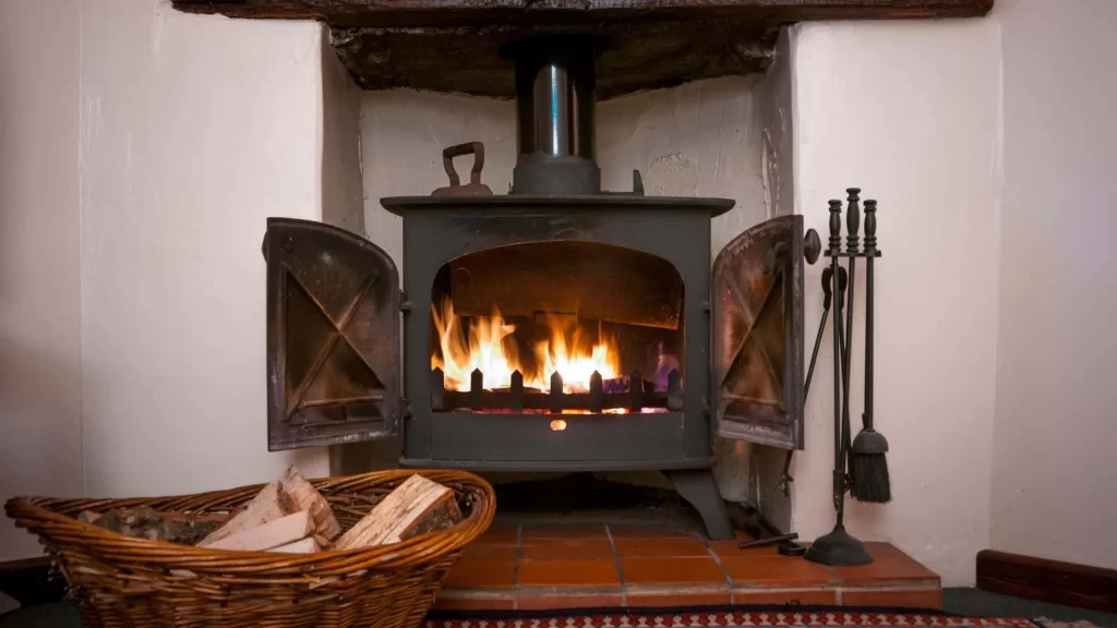 A lit wood-burning stove with open doors sits in a fireplace, with a basket of firewood on the floor nearby and fireplace tools standing to the side, creating a cozy scene much like homes cared for by air duct services New Jersey.