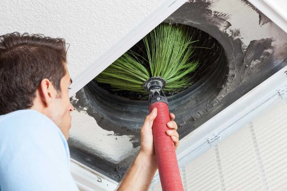 A man uses a large brush attached to a hose to clean a ceiling air duct, removing dust and debris from inside the vent&mdash;demonstrating professional air duct services New Jersey residents trust for cleaner indoor air.