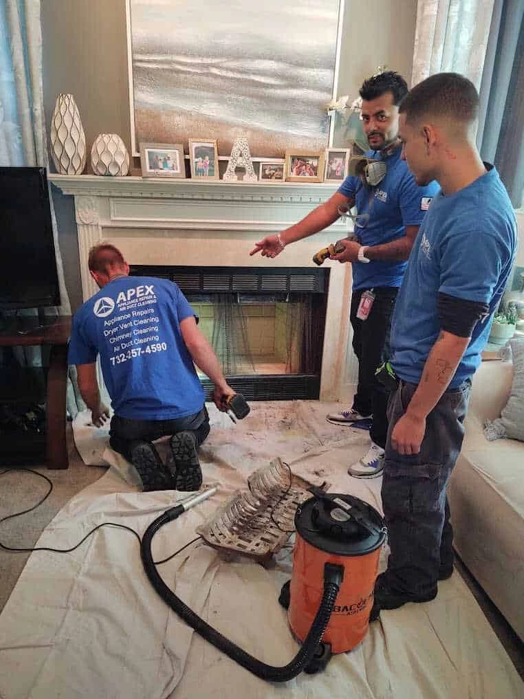 Three workers in blue Apex shirts clean a fireplace in a living room. While one kneels to work and another holds a tool, the team also specializes in air duct services New Jersey, ensuring your home stays safe and clean.