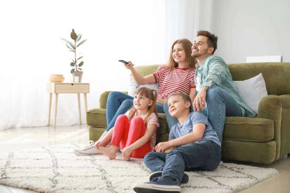 A happy family of four enjoys a bright living room, thanks to clean air from expert air duct services New Jersey, with two kids on the floor and two adults smiling on the green couch as they watch TV together.
