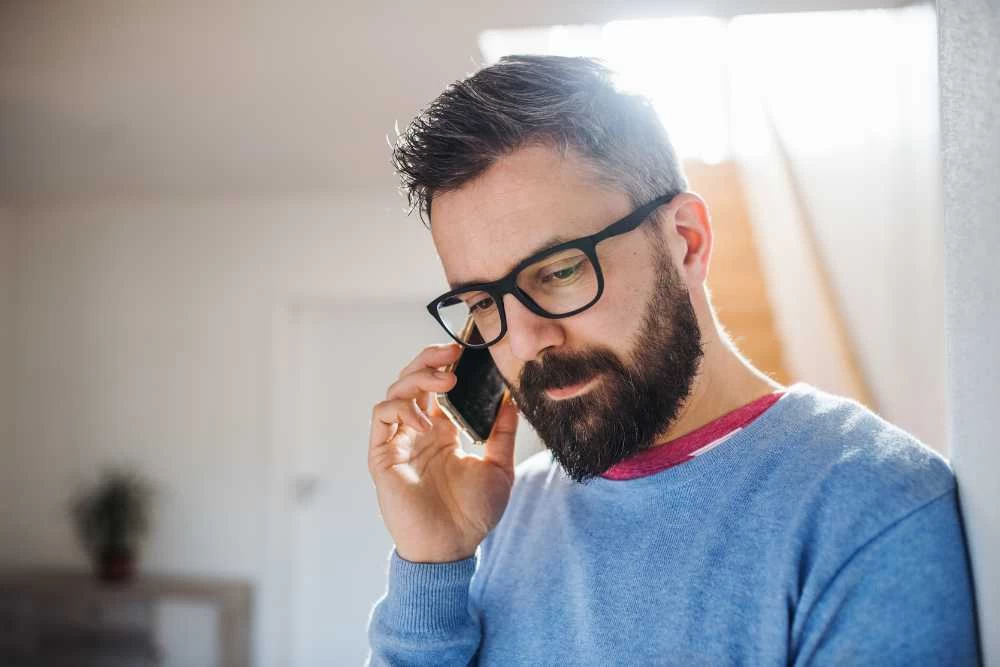 A bearded man wearing glasses and a blue sweater is standing indoors, holding a smartphone to his ear and looking down thoughtfully while talking about air duct services New Jersey. Sunlight shines in from behind him.