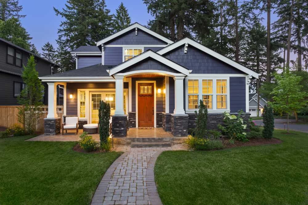 A two-story blue house with white trim, a wooden front door, porch lights on, and a landscaped yard with a curved stone path&mdash;perfect for relaxing evenings after expert air duct services New Jersey keeps your indoor air fresh and clean.