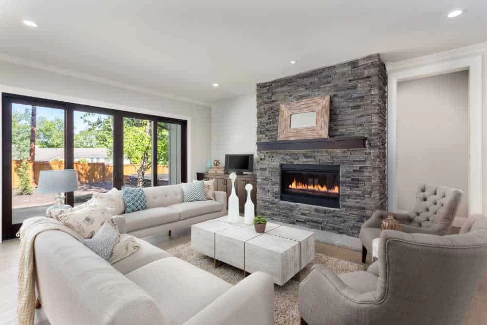 Modern living room with light-colored sofas, gray armchairs, a stone fireplace with a fire burning, a square coffee table, and large sliding glass doors overlooking a green backyard&mdash;perfect comfort after air duct services New Jersey.