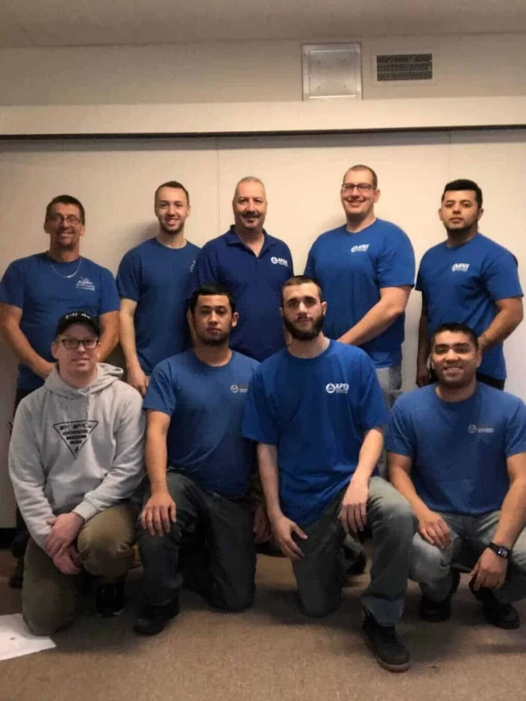 Nine men, most in blue shirts and work pants, pose indoors for a group photo. One man in front wears a gray hoodie and cap. They&rsquo;re smiling in a casual, well-lit room&mdash;your trusted team for air duct services New Jersey relies on.