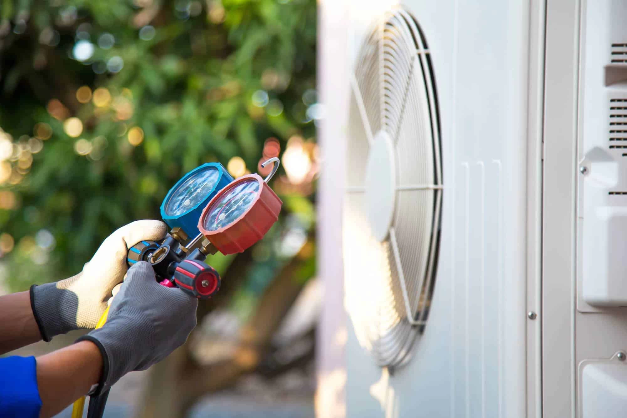A technician wearing gloves uses a manifold gauge to check or service an outdoor air conditioning unit, with green foliage blurred in the background&mdash;part of comprehensive air duct services New Jersey homeowners trust.