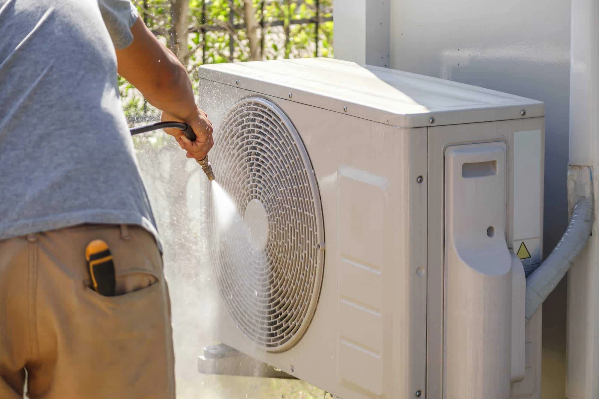 A person cleaning the outdoor unit of an air conditioner with a water hose, showcasing their attention to detail&mdash;just like with professional air duct services New Jersey relies on. The unit&rsquo;s fan grill is visible; the person wears a gray shirt and tan shorts.