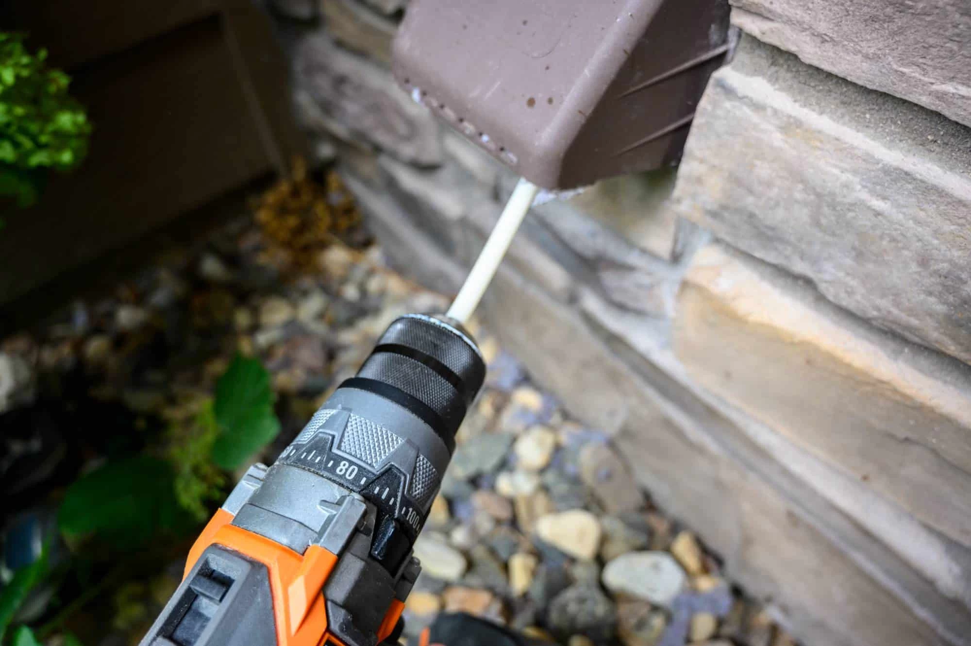 A close-up of an orange and black power drill being used to drill into the side of a house with stone siding, near a brown outdoor electrical box&mdash;typical during dryer vent cleaning New Jersey homeowners often need&mdash;surrounded by rocks and plants.