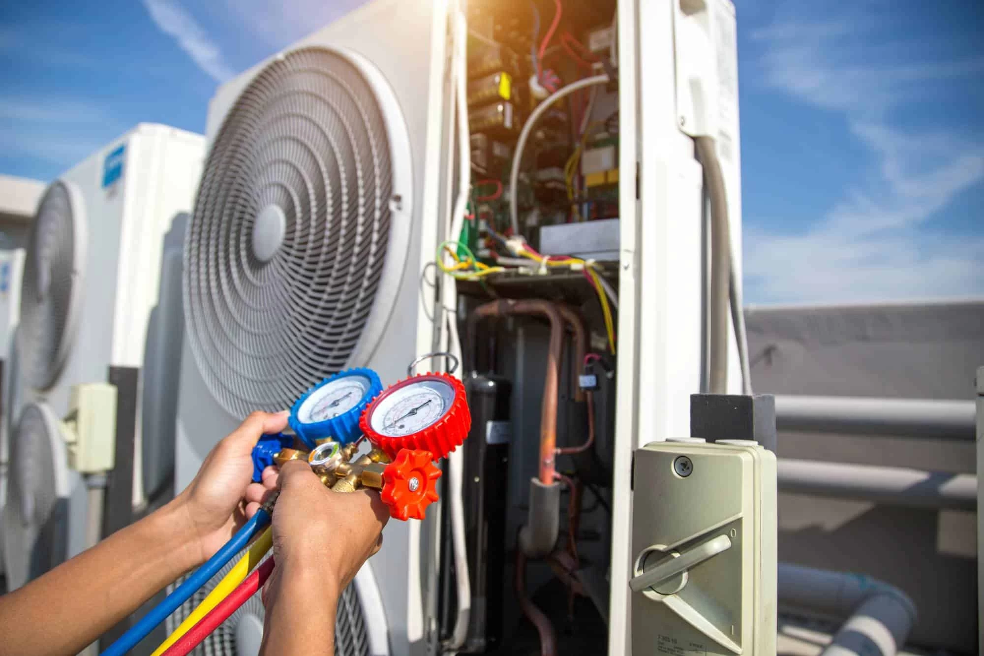 A technician uses a manifold gauge to check the pressure of an outdoor air conditioning unit, with the panel open to expose wires and components&mdash;showing the detailed care found in professional air duct services New Jersey trusts under a blue sky.