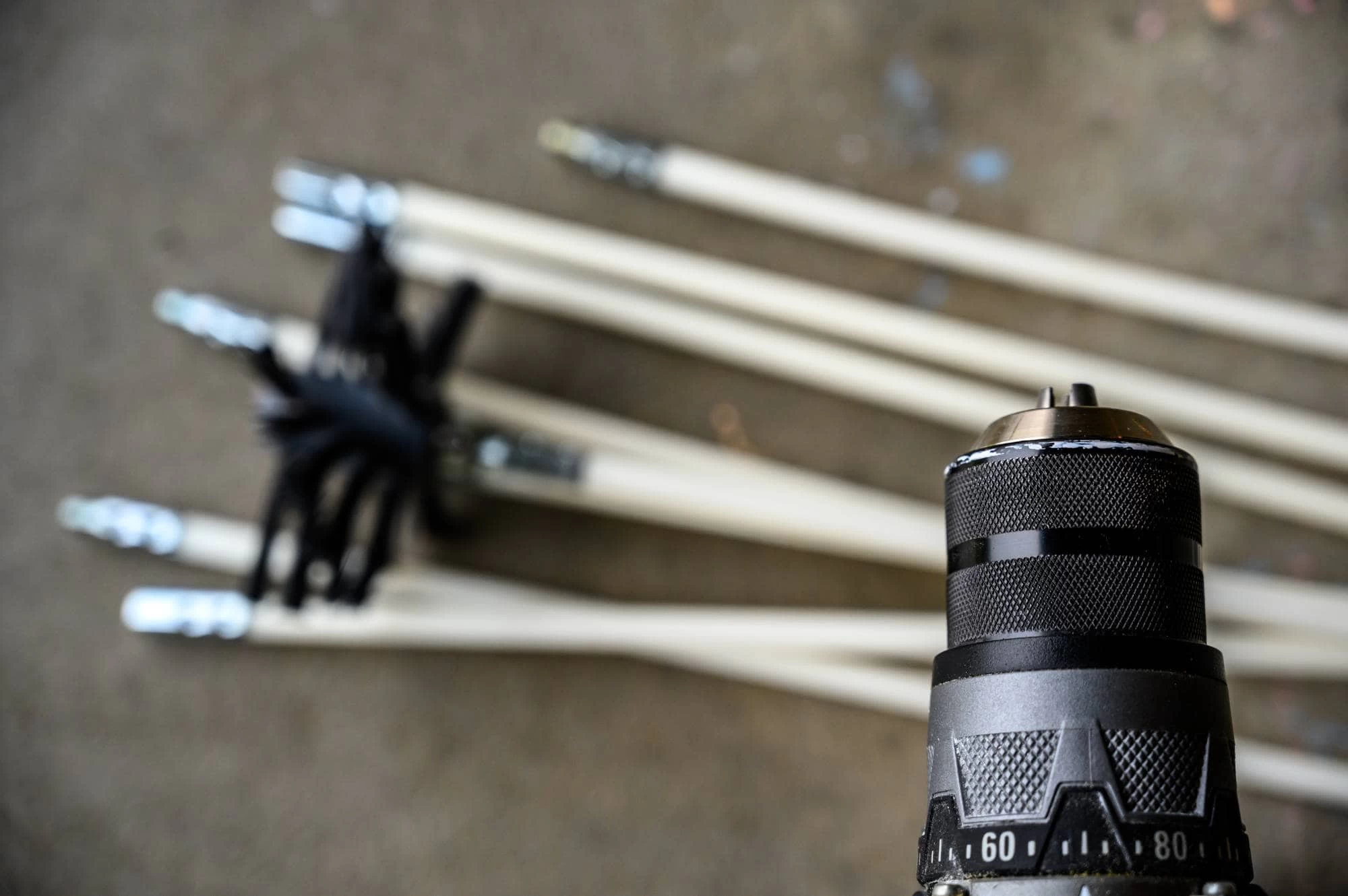 Close-up of a black and silver drill in the foreground, with several long wooden dowels with metal ends blurred in the background&mdash;tools commonly used for air duct services New Jersey on a brown surface.