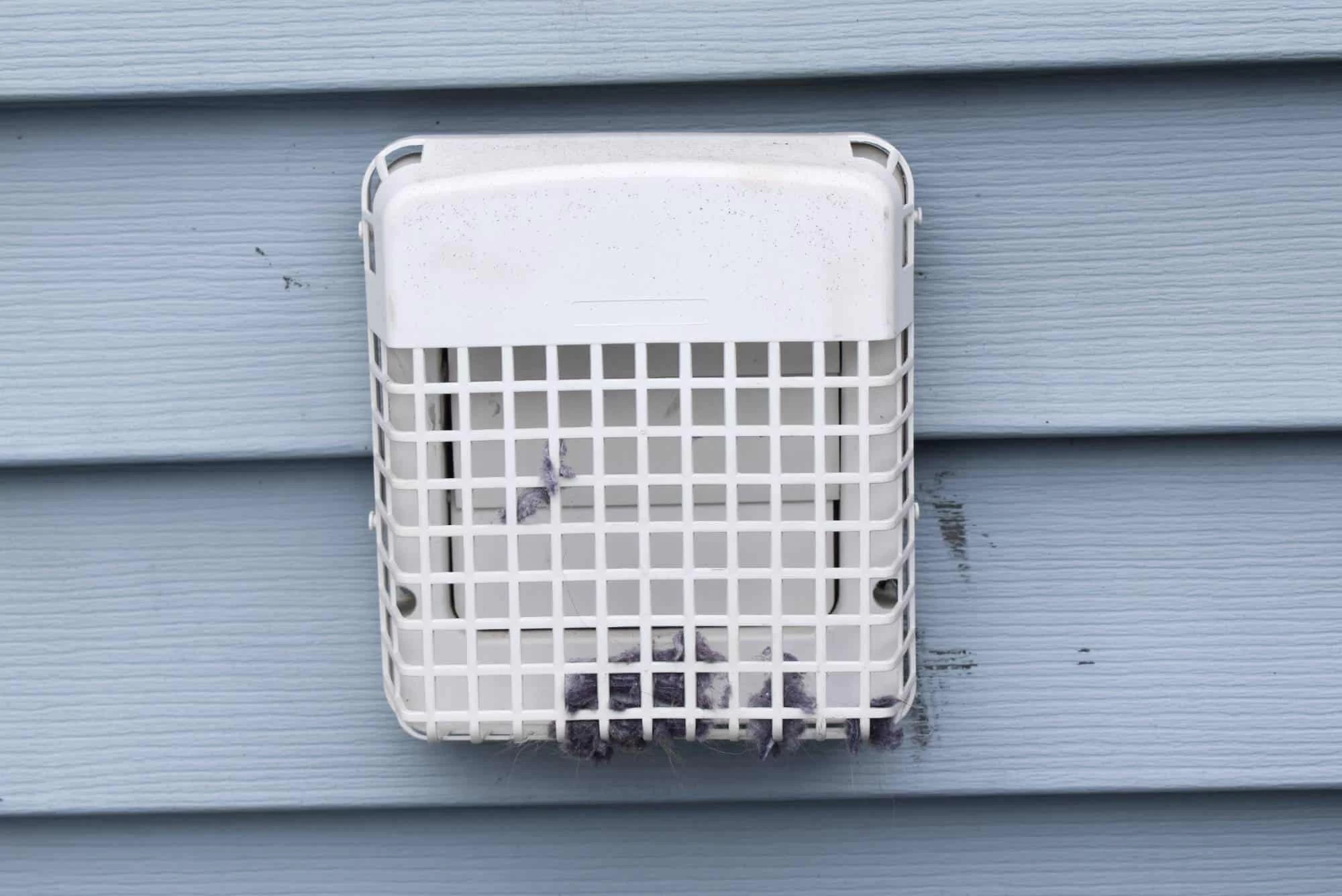A white outdoor vent cover with a protective cage is mounted on light blue siding. Some lint and debris are caught at the bottom of the cage—a clear reminder of the importance of regular dryer vent cleaning New Jersey homeowners need.