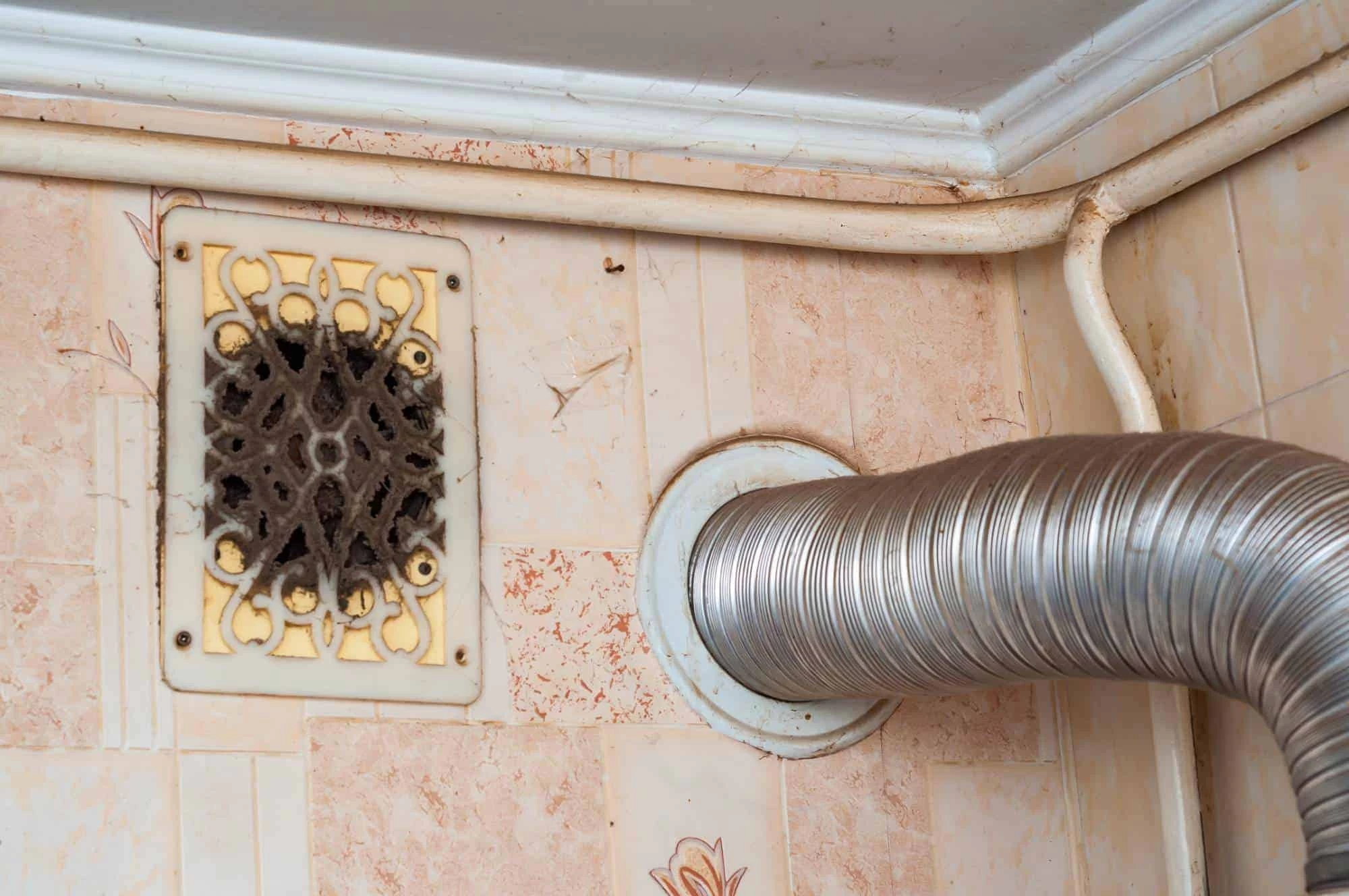 A dusty, ornate vent cover and a metal duct pipe installed in the corner of a room with tiled walls and white molding&mdash;perfect examples of areas that benefit from professional air duct services New Jersey.