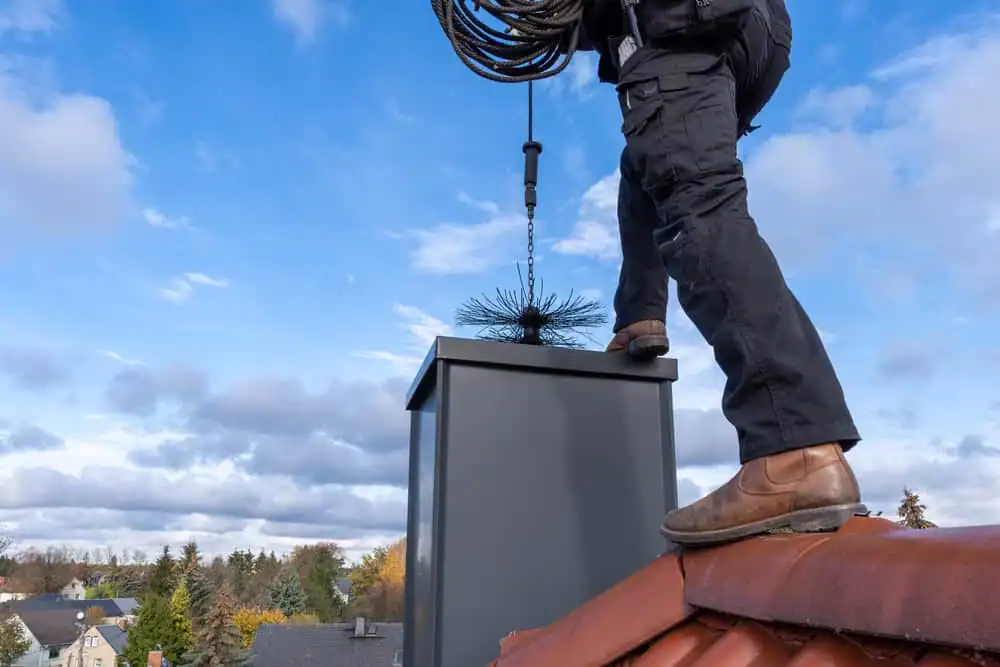 Person wearing black pants and brown shoes stands on a red tiled roof, cleaning a chimney with a brush attached to a long flexible rod&mdash;similar to the meticulous care seen in dryer vent cleaning New Jersey; trees and cloudy sky are visible in the background.