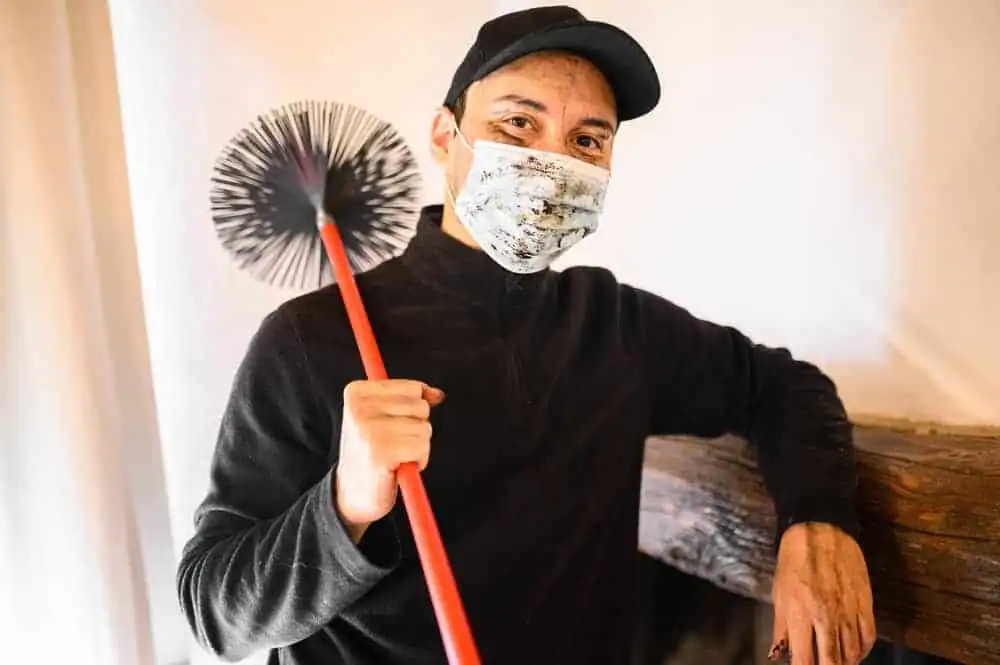 A person wearing a black cap, patterned face mask, and dark sweater holds a chimney sweep brush with a red handle while standing next to a wooden beam indoors&mdash;perfectly suited for dryer vent cleaning New Jersey.