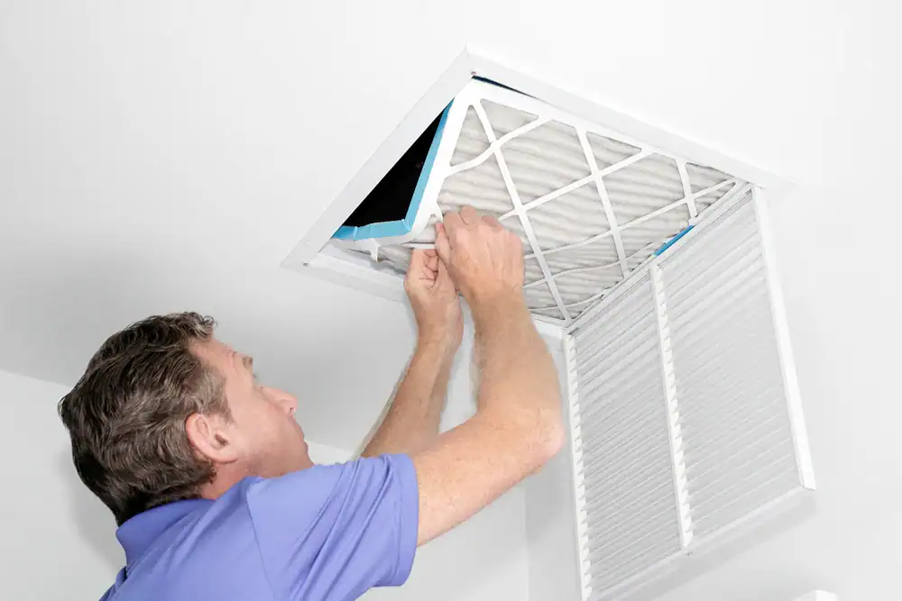A man in a blue shirt is replacing or installing an air filter in a ceiling vent, with the vent cover open&mdash;part of routine air duct services New Jersey homeowners trust for healthier indoor air.