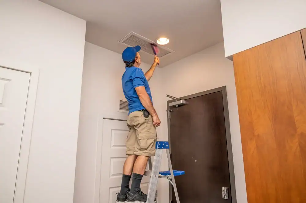 A person in a blue shirt and cap stands on a step ladder, cleaning an air vent on the ceiling with a pink cloth. The room has white walls and a wooden door, highlighting the importance of regular air duct services New Jersey.