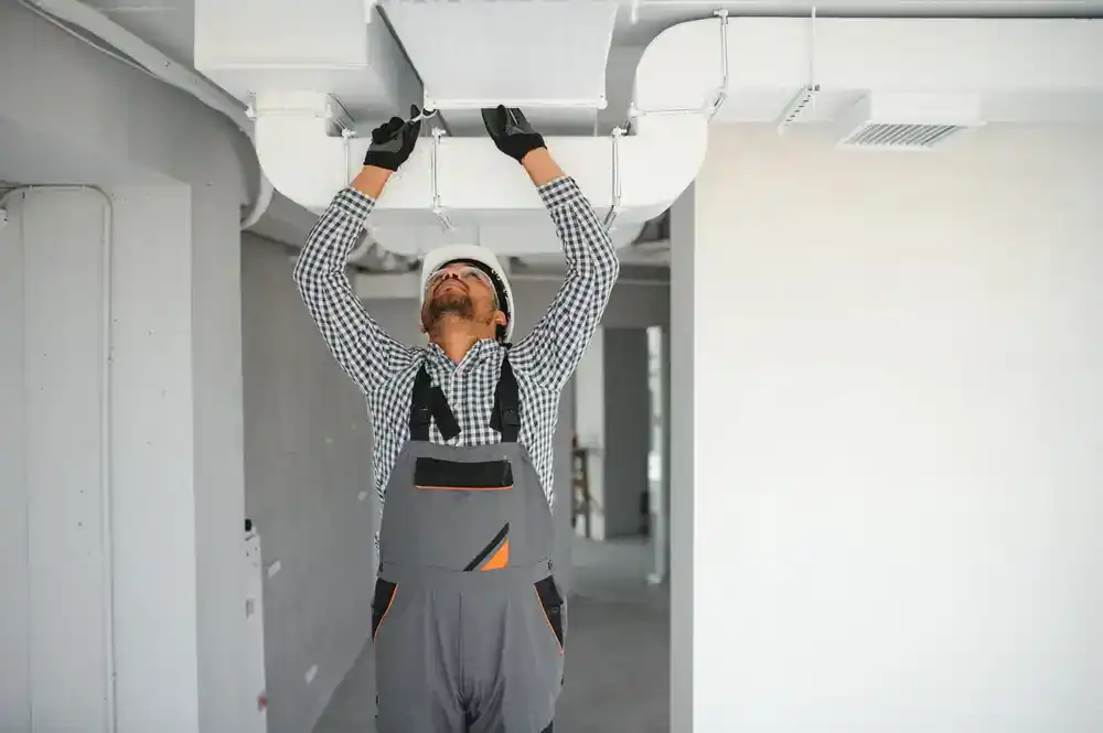 A construction worker in overalls, gloves, and safety glasses installs or repairs an air duct on the ceiling inside a building, providing expert air duct services New Jersey residents can trust.