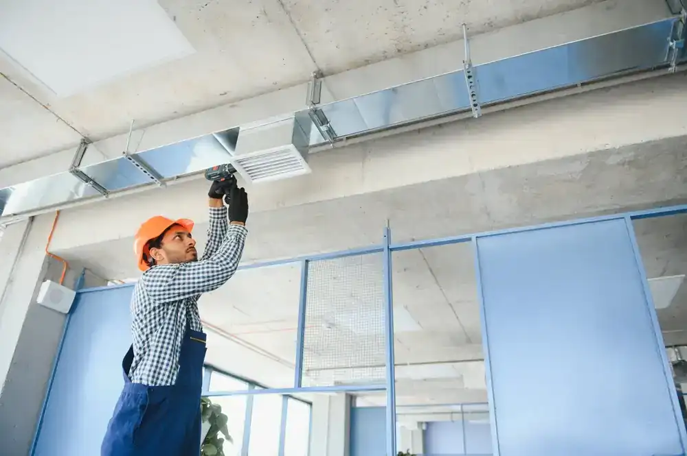 A worker in a hard hat and gloves uses a power drill to install or repair an air vent on the ceiling, showcasing professional air duct services New Jersey inside a modern office building with glass partitions.