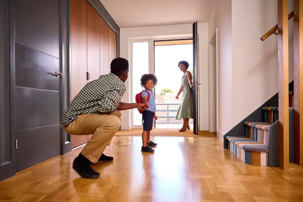 A man kneels on the floor, holding a young child's hand in a bright hallway. A woman stands by the open front door, smiling at them. Stairs are visible on the right, and sunlight streams in&mdash;perfect for families seeking dryer vent cleaning New Jersey.
