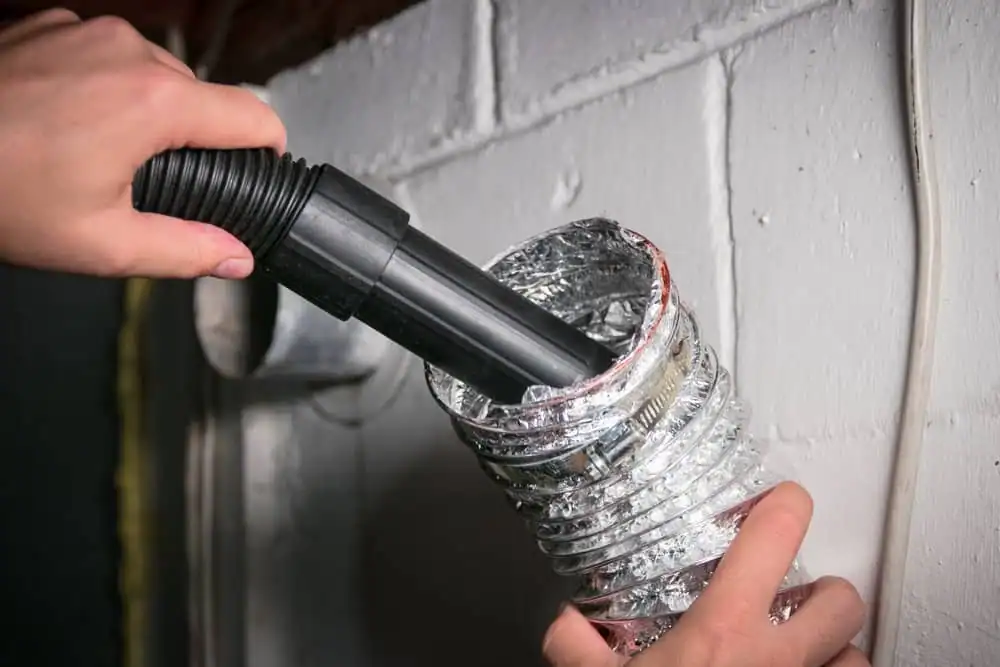 A person uses a vacuum hose to clean the inside of a flexible metal dryer vent duct attached to a white wall, demonstrating professional dryer vent cleaning New Jersey homeowners can trust.
