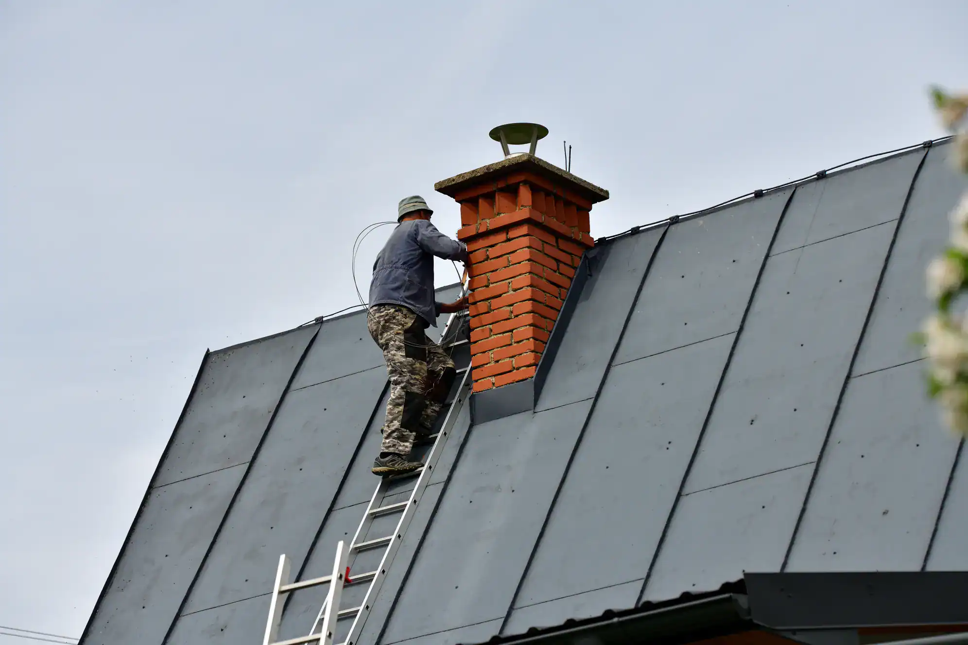 A person in camouflage pants and a bucket hat stands on a ladder, cleaning or inspecting a red brick chimney on a gray metal roof of a building.