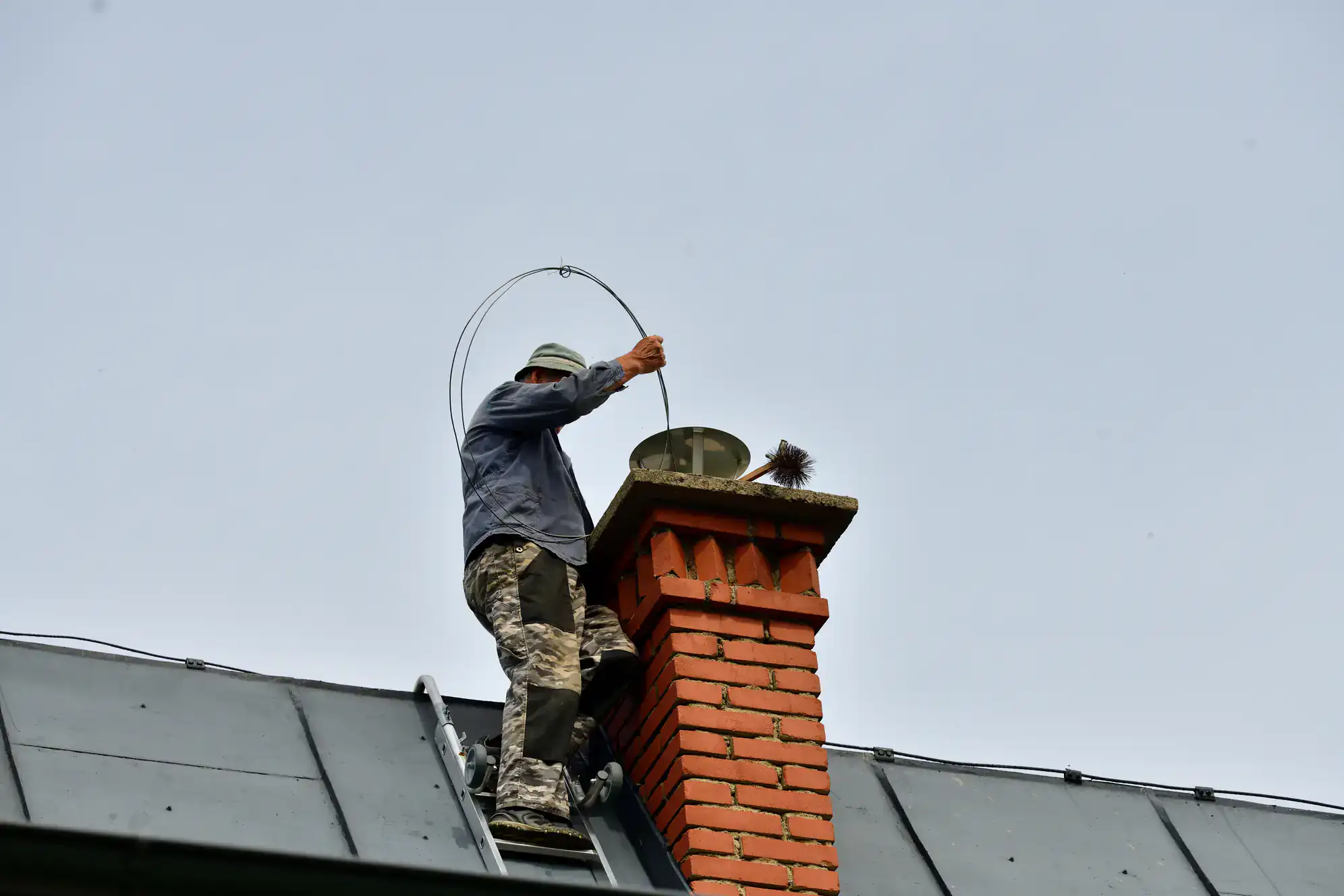 A person standing on a ladder by a brick chimney, cleaning it with a long flexible brush, wearing a hat and camouflage pants, under a clear sky.