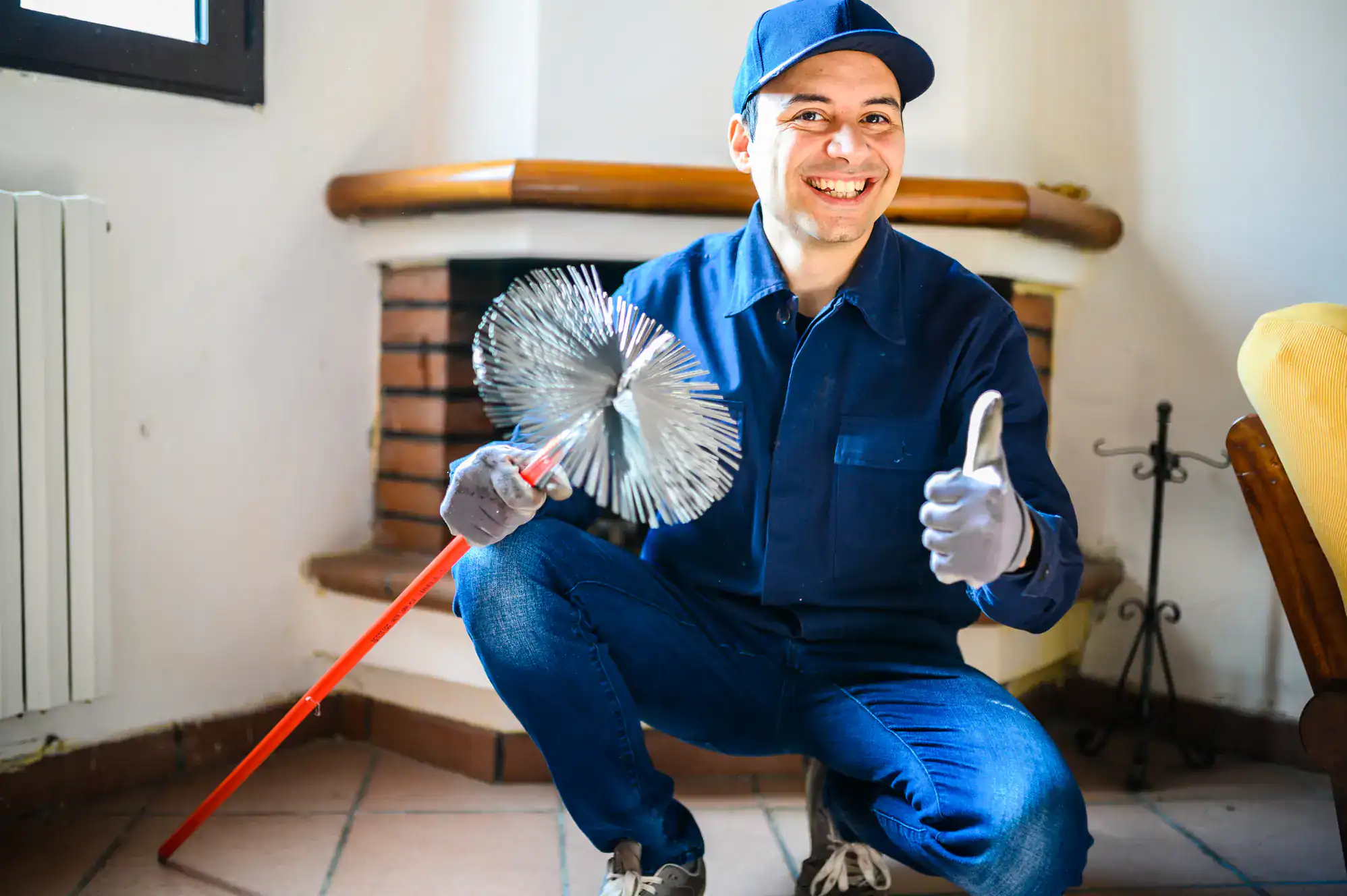 A smiling man in blue work clothes and cap kneels indoors, holding a chimney cleaning brush and giving a thumbs-up gesture. He appears to be a chimney sweep in a living room with a fireplace in the background.