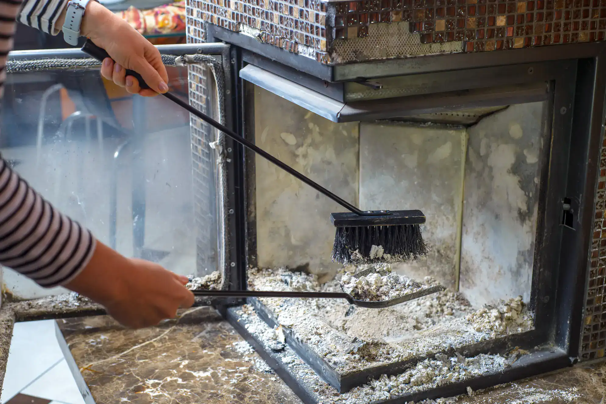 A person in a striped shirt uses a brush and dustpan to clean ash from the inside of a fireplace with glass doors and a mosaic tile exterior.