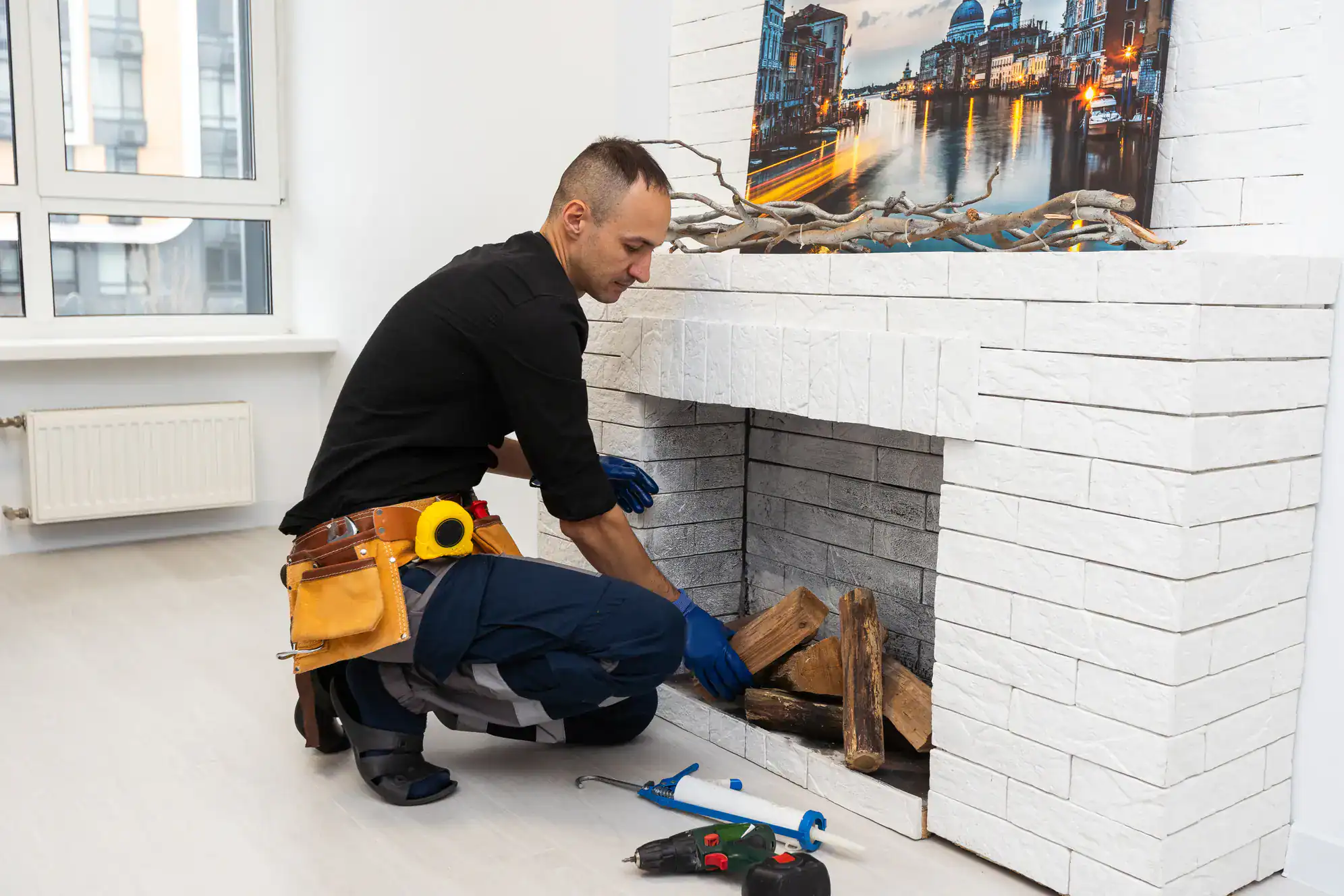 A man wearing a tool belt and gloves arranges firewood inside a white brick fireplace in a modern room. Tools are placed on the floor nearby, and a cityscape photo decorates the mantel above.