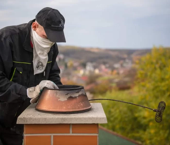 A person in work clothes and a cap, with a cloth covering their face, cleans a chimney on a rooftop using a long brush&mdash;highlighting the importance of air duct services New Jersey for maintaining safe and efficient home environments.
